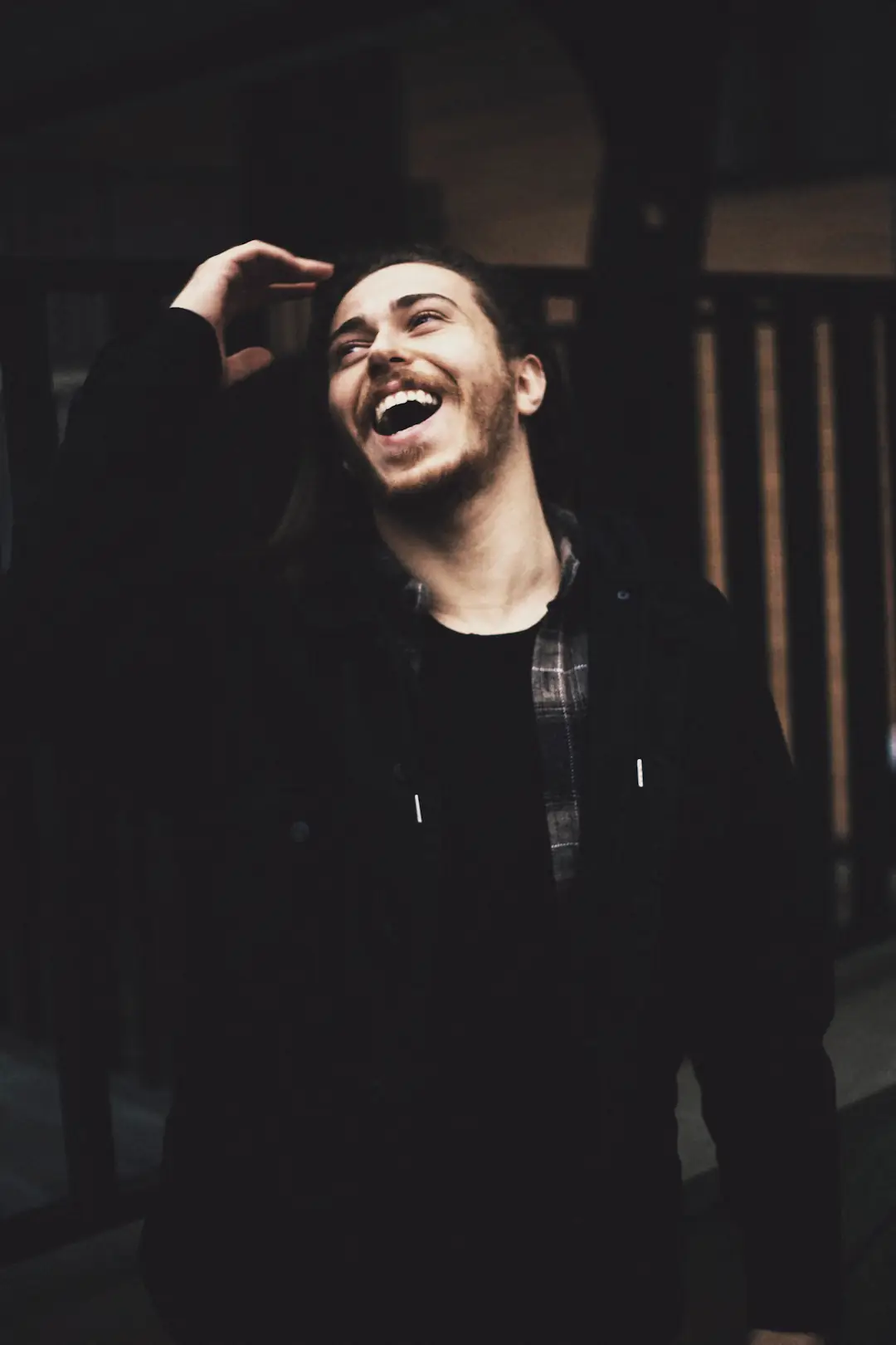 A young man in a dark jacket laughing, head back, a hand in his hair.
