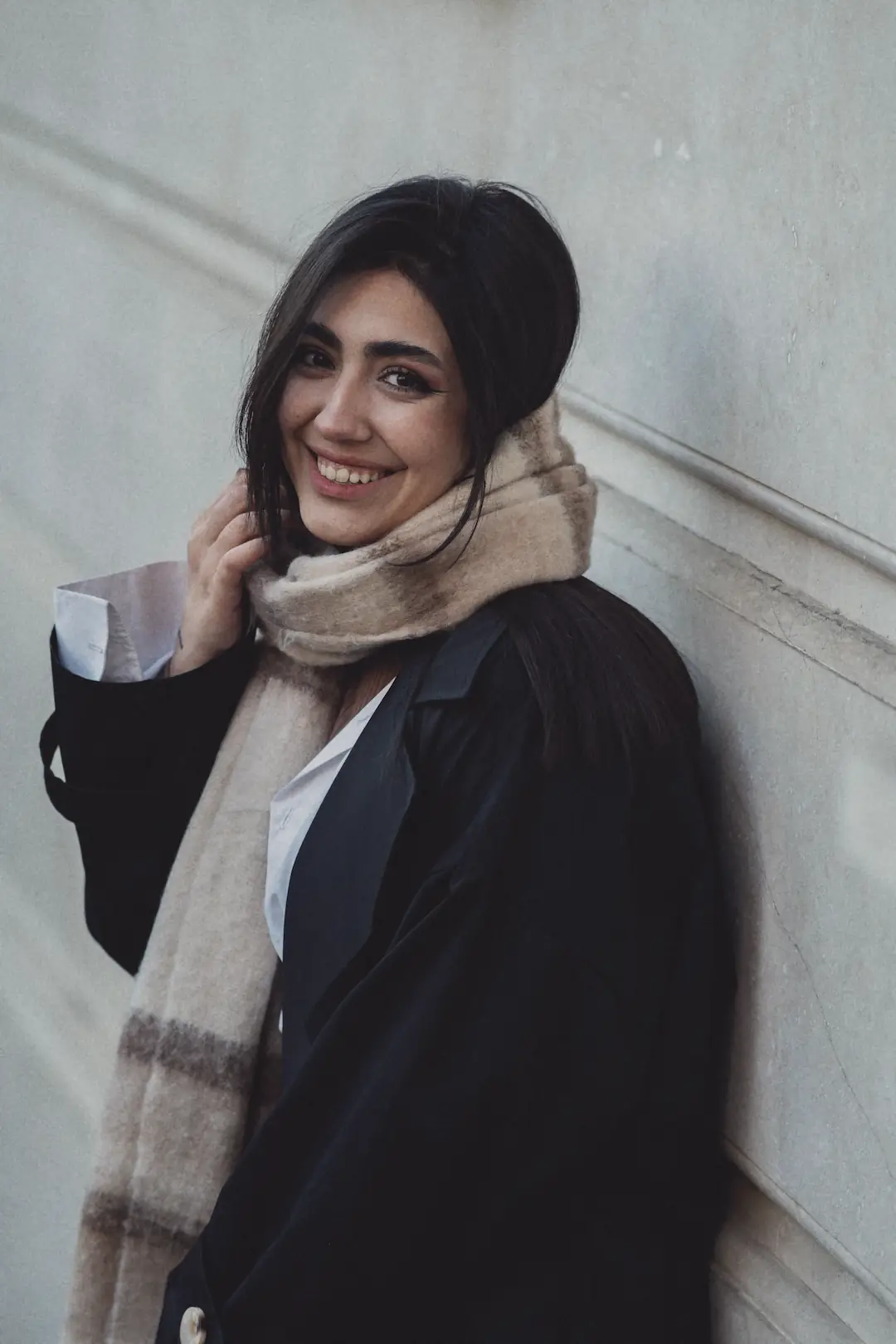 A smiling woman in a black coat and check scarf leaning on a pale wall.